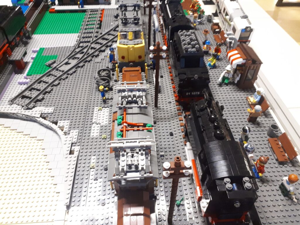 LEGO treinbaan met grijze 9V-rails, klassieke locomotief en wagons in aanbouw op een grote groene plaat